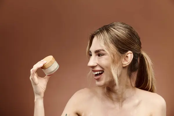 stock image A young woman with vitiligo and a tattoo smiles while holding a jar of cream against a beige background.