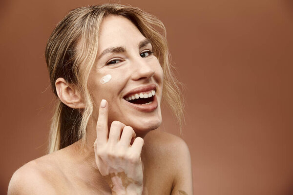 Молодая женщина с витилиго улыбается, когда она наносит крем на лицо на бежевом фоне.