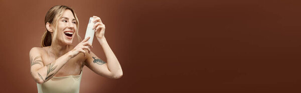 Молодая женщина с витилиго улыбается, показывая продукт по уходу за кожей на бежевом фоне. Она носит случайный топ и татуировку на руке..