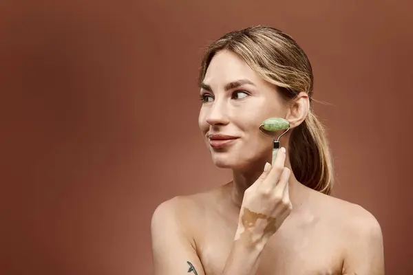 stock image A young woman with vitiligo uses a jade roller on her face as part of her skincare routine. The background is a soft beige.