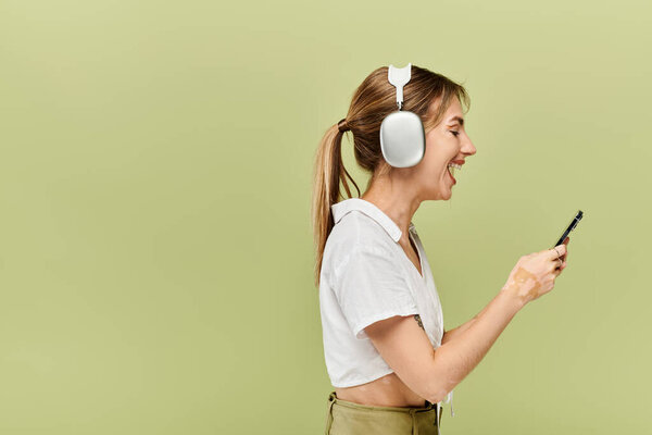 A young woman with vitiligo smiles and looks at her phone while wearing a white summer outfit and headphones against a green background.