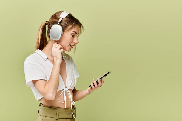 A young woman in white crop top and khaki pants poses against green backdrop, with headphones on and looking at phone.