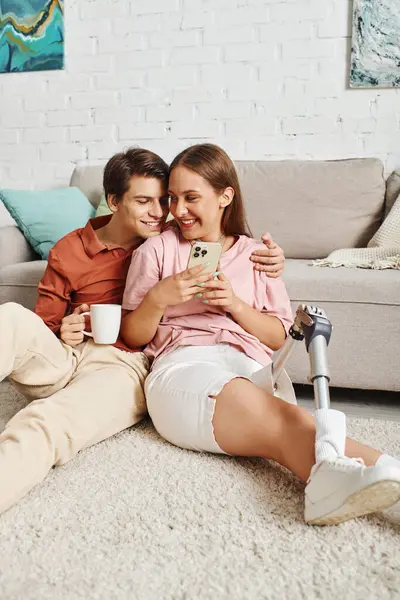 stock image A couple sits on the floor, looking at a phone together, while enjoying a cup of coffee.