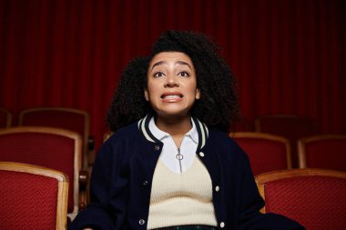 A young woman in stylish casual clothes sits in a movie theater, reacting with surprise and wide eyes to the screen. clipart