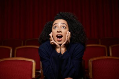 Sinemadaki genç bir Afrikalı Amerikalı kadın şaşkınlıkla tepki veriyor, elleri ağzını kapatıyor..