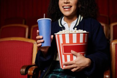 A young woman watches a movie in a cinema, holding a large bucket of popcorn and a beverage. She appears captivated by the film. clipart