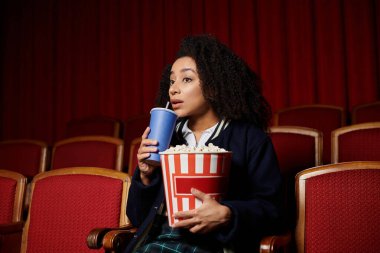A young woman with curly hair watches a movie with popcorn and a drink, captivated by the screen.