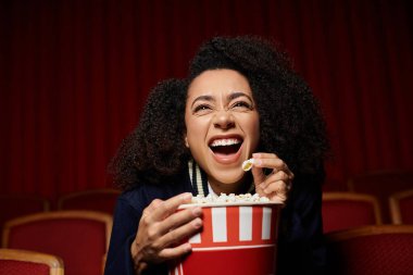 Kıvırcık saçlı genç bir kadın elinde patlamış mısır kovasıyla karanlık bir sinemada film izlerken neşeyle gülüyor..
