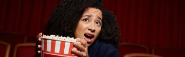 Curly-haired woman in theater seat, clutching popcorn, looks shocked and scared at the movie screen. clipart