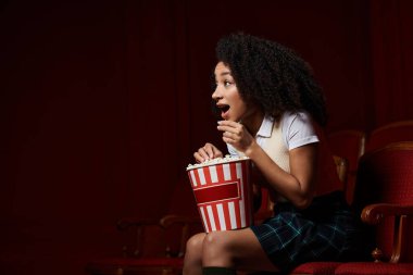 A young woman, dressed casually, sits in a cinema seat, watching a movie with an excited expression, holding a bucket of popcorn.