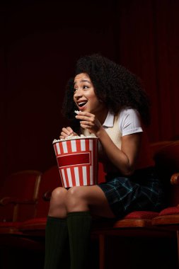 Sinema koltuğundaki kıvırcık saçlı siyah bir kadın elinde patlamış mısır tutarken heyecanla tepki veriyor..