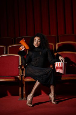 A fashionable African American woman in an elegant black dress sits in a red cinema seat, reacting to the movie with a megaphone.