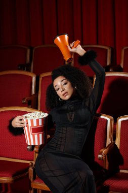 A stylish woman enjoys a movie in a vintage theater, holding popcorn and a megaphone. clipart