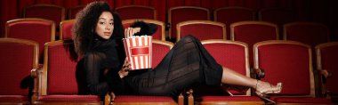 An elegantly dressed African American woman watches a movie in a theater, visibly enjoying the film and a bucket of popcorn.