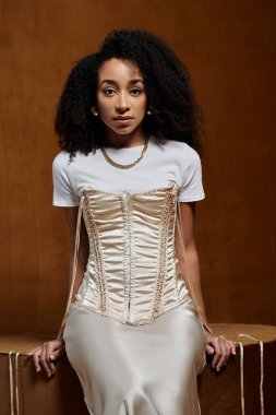 Beyaz bluz ve korse giyen şık bir Afro-Amerikan kadın stüdyo ortamında kendine güvenerek poz veriyor..