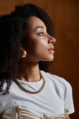 A fashionable African American woman with curly hair poses in a studio, wearing a gold chain necklace and earrings. clipart