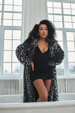 Kıvırcık saçlı, leopar desenli ceket ve siyah elbise giyen şık bir Afrikalı Amerikalı kadın.