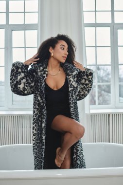 Moda desenli leopar desenli ceketli şık bir Afro-Amerikalı kadın stüdyo ortamında, küvette poz veriyor..