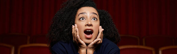 A young Black woman with curly hair sits in a movie theater, hands covering her mouth in surprise as she watches the film.