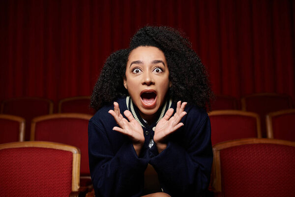 Черная женщина в повседневной одежде сидит в кинотеатре, руки подняты в шоке на сцене на экране.