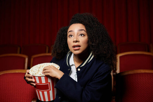 Молодая афроамериканка в стильной повседневной одежде смотрит кино в кинотеатре с удивлением и волнением..