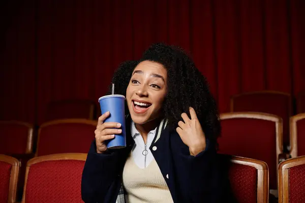 stock image A young woman in a cinema seat reacts enthusiastically to a movie, holding a drink and smiling widely.