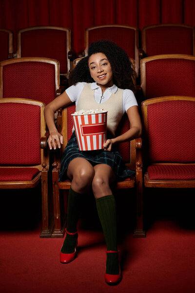 Молодая женщина с вьющимися волосами сидит на сиденье кинотеатра, держа ведро попкорна, во время просмотра фильма.