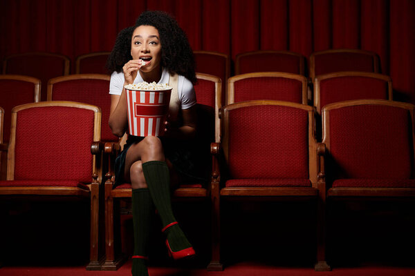 Молодая женщина, одетая в стильную повседневную одежду, смотрит кино в кинотеатре и реагирует на экран, наслаждаясь закуской.