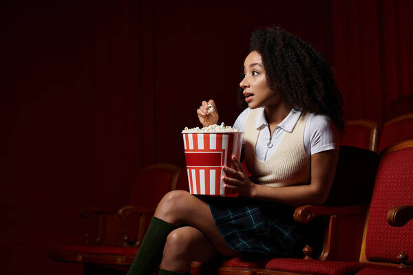 A young woman captivated by the film on screen, sits in a theater seat with a bucket of popcorn, eagerly watching.