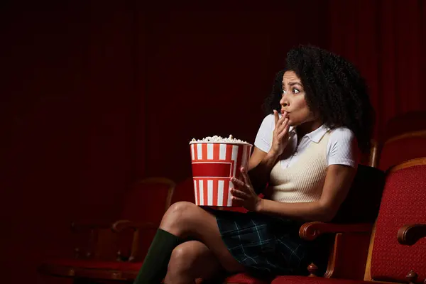 stock image A young African American woman sits in a movie theater, engrossed in the film, popcorn in hand.