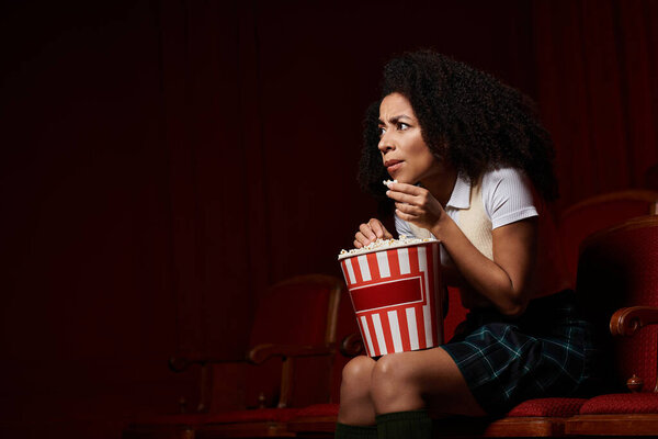 A young woman sits in a theater seat, engrossed in a movie, with a look of anticipation on her face.