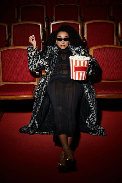 Стильная афроамериканка в леопардовом пальто смотрит фильм в темном кинотеатре.