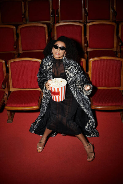 Стильная женщина в леопардовом пальто смотрит фильм, держит попкорн и реагирует на фильм.