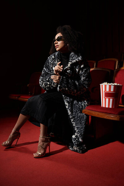 A stylish African American woman, dressed in elegant attire, enjoys a movie in a dark cinema.