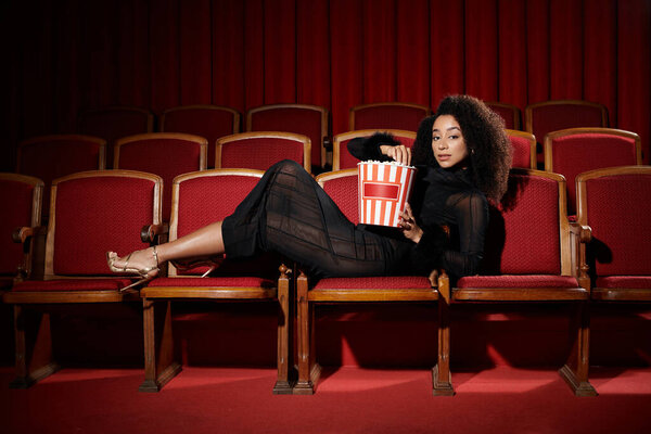 Стильная афроамериканка в черном платье наслаждается фильмом в кинотеатре, держа ведро попкорна и реагируя на фильм.