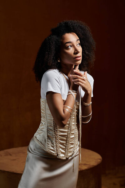 A stylish African American woman with curly hair poses in a studio setting, wearing a white top and a fitted corset.