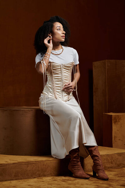 A woman in a white top, silky dress with fitted bodice sits on a platform. She has dark, curly hair and brown boots.