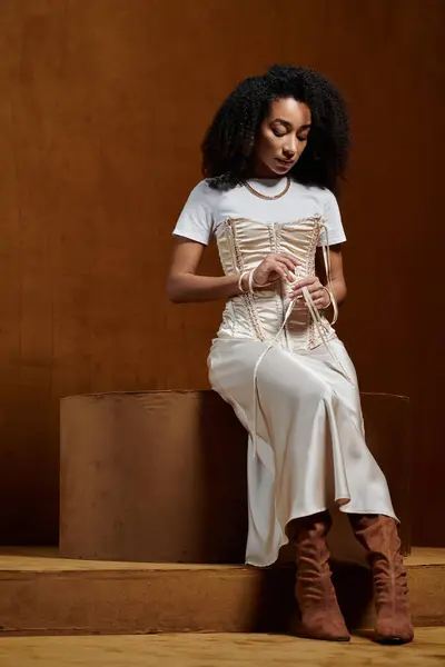 stock image An African American woman in a white top and a cream satin skirt poses in a studio setting, adjusting the corset.