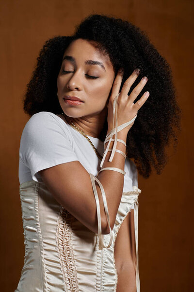 В студии шикарная афроамериканка надевает белый топ и кремовый корсет, глаза закрыты, рука отдыхает на лице.