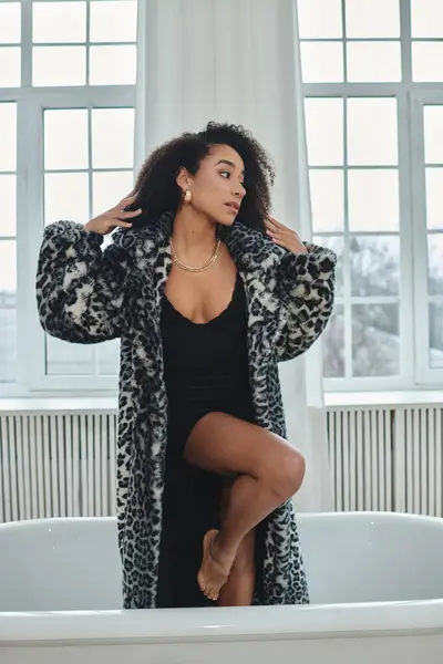 stock image A stylish African American woman in a trendy leopard print coat poses in a studio setting, standing in a bathtub.