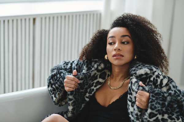 A young woman with curly hair poses in a studio wearing a leopard print coat and black top.