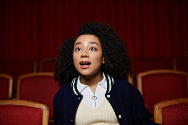 A young woman in a dark blue jacket sits in a movie theater, watching the screen with a shocked expression.
