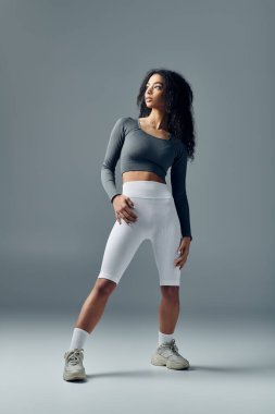 Gri bluzlu ve beyaz şortlu genç bir kadın, gri bir arkaplana karşı güvenle poz veriyor..