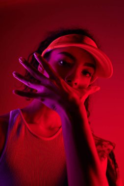 A young woman in a tank top and visor poses dramatically against a vibrant red backdrop illuminated by neon lights. clipart