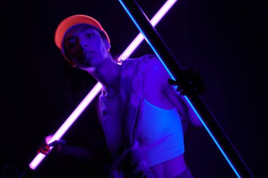 Parlak turuncu şapkalı genç bir kadın stüdyoda poz veriyor, neon ışıklarıyla yıkanmış..