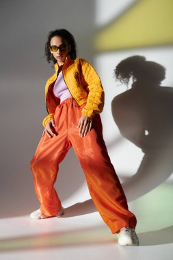 A stylish young woman poses confidently in a yellow jacket and orange pants, her sunglasses adding an edge to her look. clipart