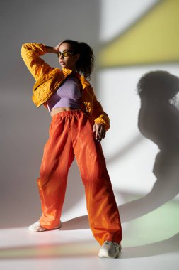 Stüdyo ortamında sarı ceketli ve turuncu pantolonlu şık bir Afrikalı Amerikalı kadın poz veriyor..