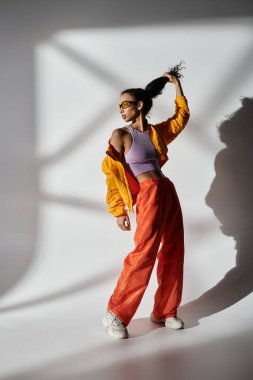 Sarı ceketli ve turuncu pantolonlu genç bir kadın stüdyo ortamında poz veriyor, güneş gözlükleri ışığı yansıtıyor..