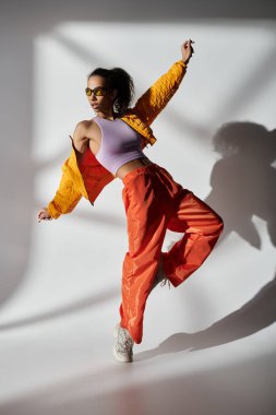 Genç bir Afrikalı Amerikalı kadın stüdyoda poz veriyor, sarı ceket, turuncu pantolon ve güneş gözlüğü giyiyor..