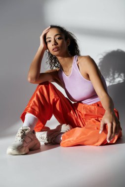 Modaya uygun turuncu pantolon ve lavantalı bluz giyen genç bir kadın gri arka planlı bir stüdyoda oturuyor..